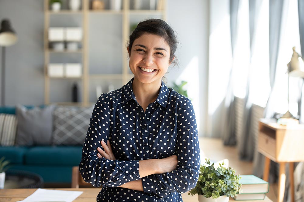 Smiling pretty young adult indian woman looking at camera posing at modern home arms crossed